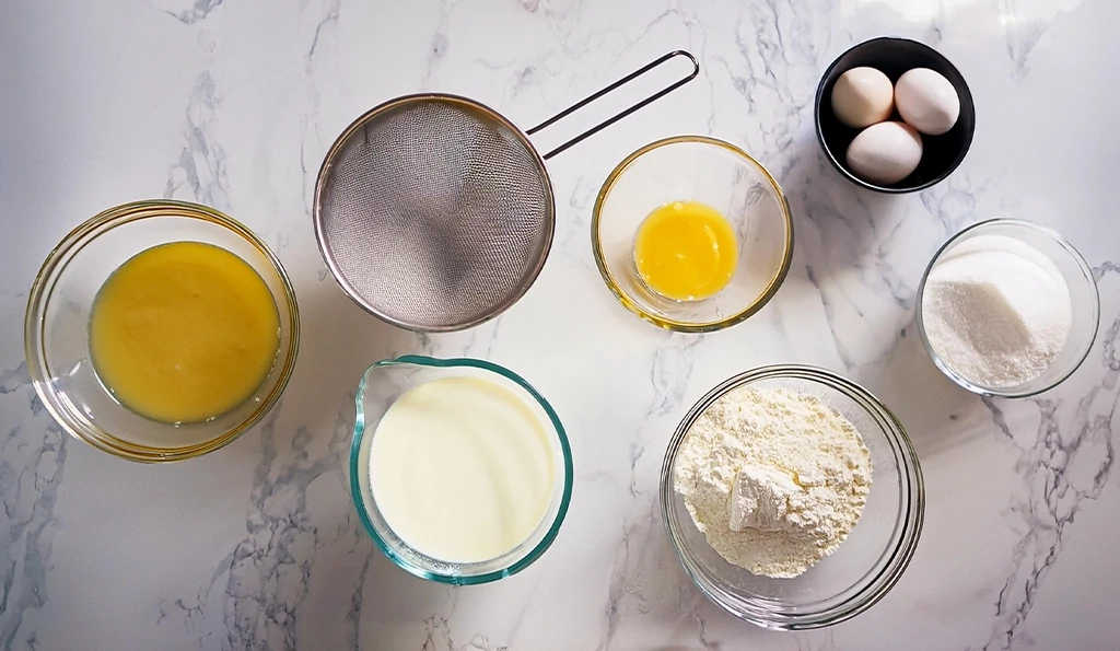 Ingredients for Hot Milk Cake Recipe arranged on a white marble countertop, including milk, butter, eggs, flour, sugar, vanilla extract, and baking powder, ready for baking.