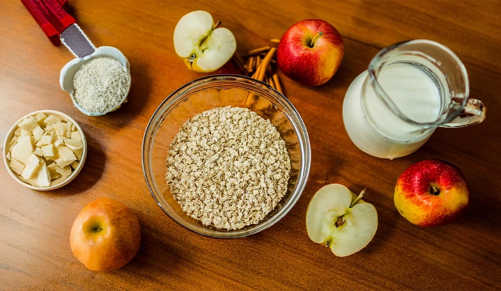 Flat-lay of Protein Baked Oatmeal ingredients, including rolled oats, protein powder, almond milk, chopped apples, cinnamon, maple syrup, and baking powder, arranged on a rustic wooden table.