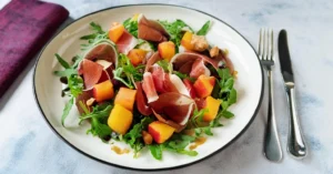 Plate of fresh melon and arugula salad topped with thin slices of Parma ham (prosciutto di Parma), drizzled with olive oil and garnished with cracked black pepper, served on a white plate with a purple napkin and silver fork.